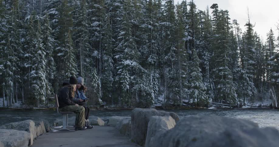 Two girls taking his selfie while sitting on a bench next to a cold river