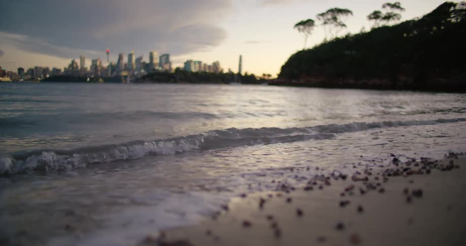 Waves roll onto sandy beach past promontory as city skyline sits on horizon under red tinted sky