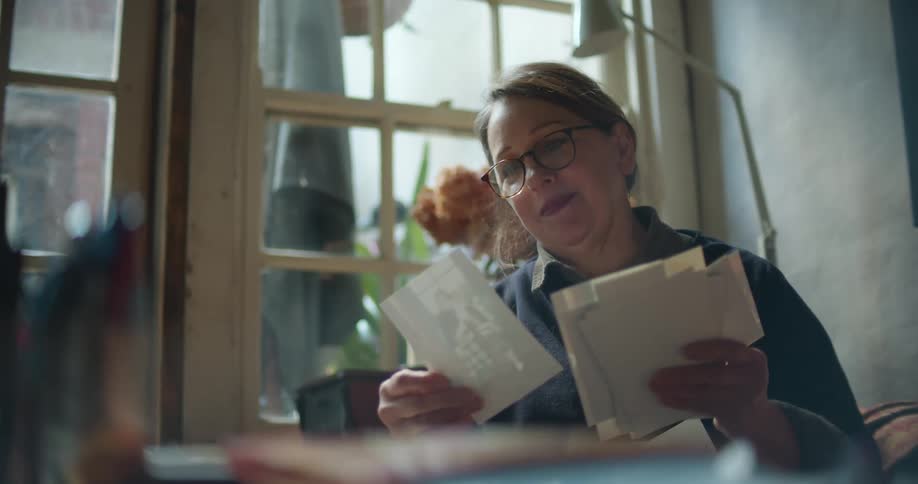 Woman wearing eyeglasses sites near window sorting through photographs and smiling
