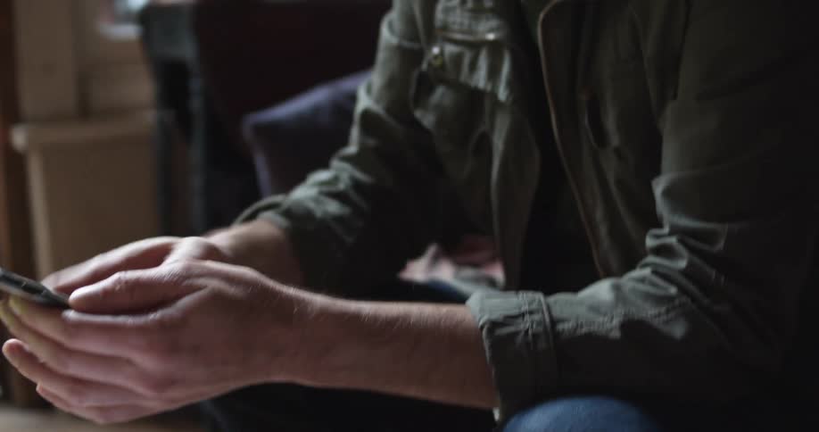 A man with headphone on sits on his couch at home and talks with someone on his laptop and then picks up his cell phone and texts