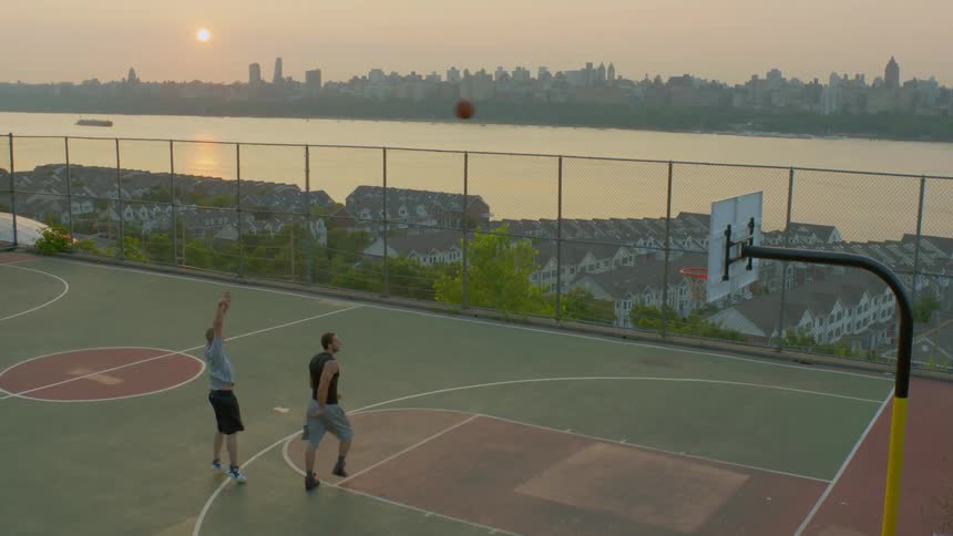 Two men play basketball on a city court at dusk