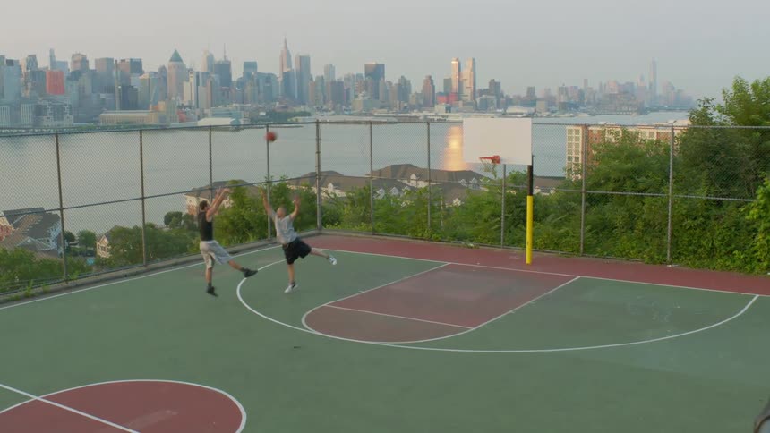 Two men play basketball on an outdoor court with a long view of a city's skyline and waterway