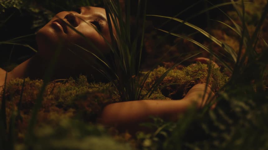 A female poses for a photo shoot by laying on some soil and surrounded by plants