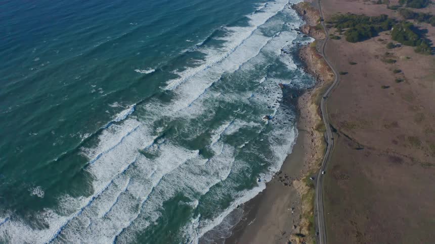 Ocean waves roll towards the beach while cars travel on the nearby highway