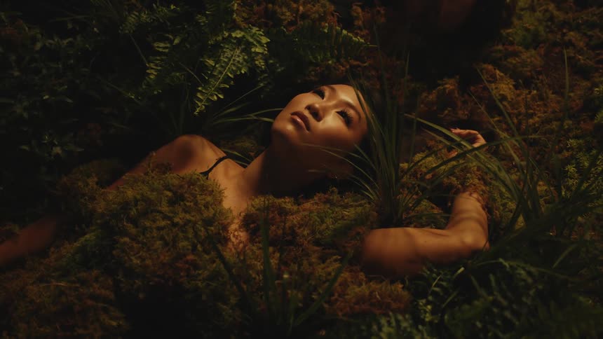 A woman lies staring up with her body is covered by moss