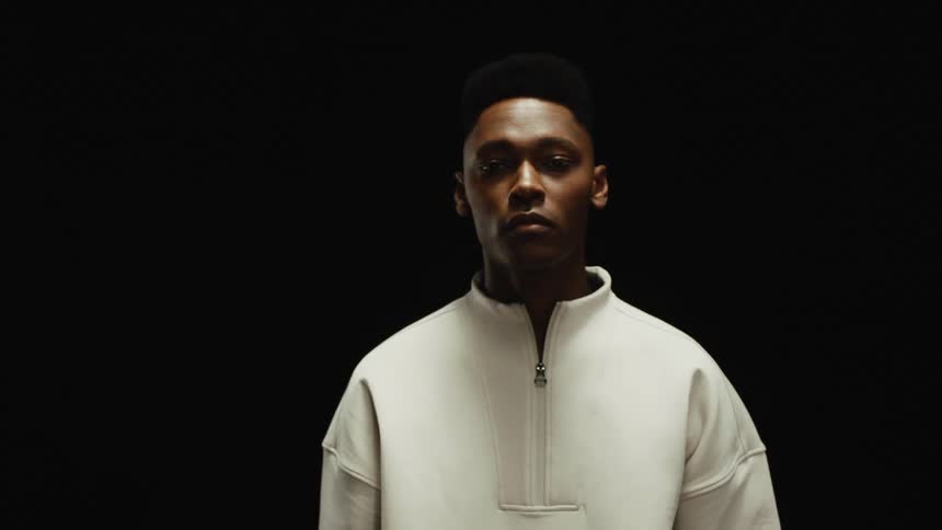Man wearing white zippered sweat shirt walks forward surrounded by darkness