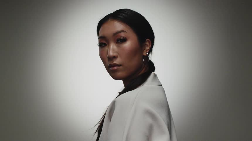 Woman with hoop earrings straight hair and a white blouse faces the camera in front of grey background