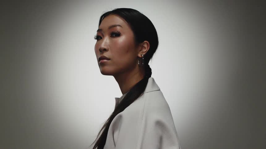 Woman with straight hair hoop earrings and a white blouse looks at the camera in front of grey background