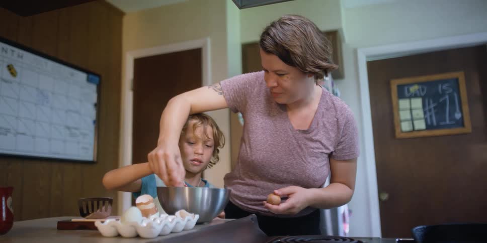 A woman and a male child mix the contents of a steel bowl next to an egg carton