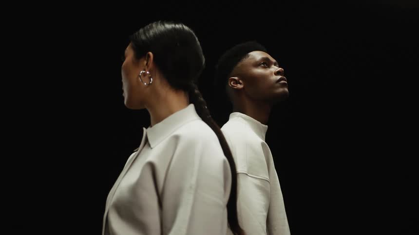 Two models in oversized white shirts stand back to back in a dark room