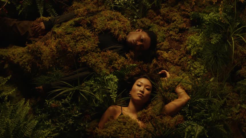 Two people are laying covered in plant material while the woman slowly moves her head