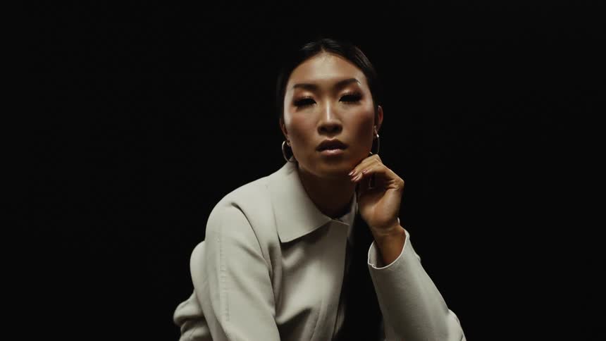 Young asian model sitting in a room in front of a black background looks at the camera