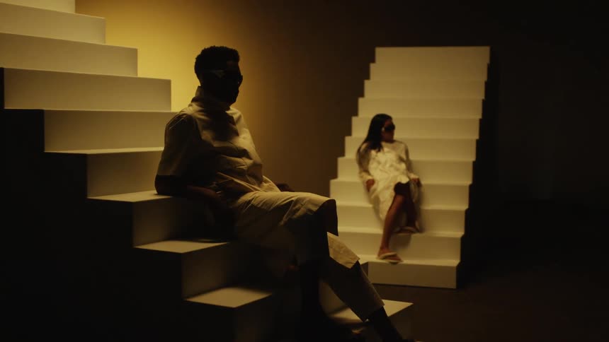 A woman and a man sitting on flights of white stairs wearing sunglasses and looking away from the camera