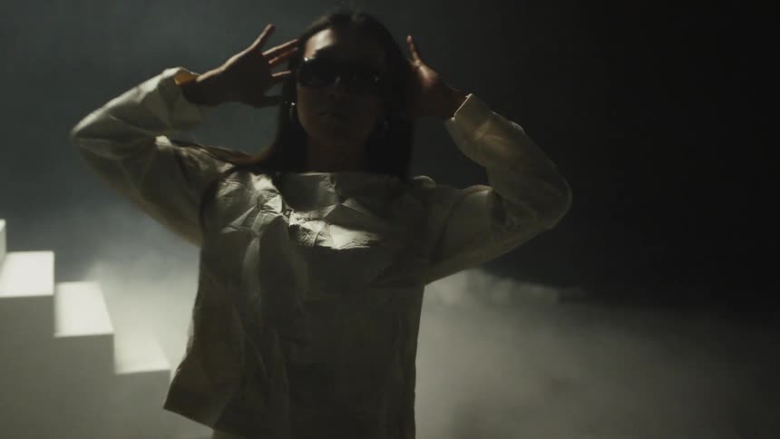 Young woman wearing sunglasses and a white dress models for the camera in a room filled with smoke