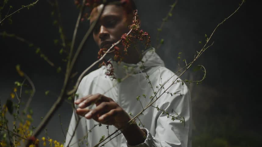 Man wearing white jacket with hood stands touching bush as vapor rises behind him