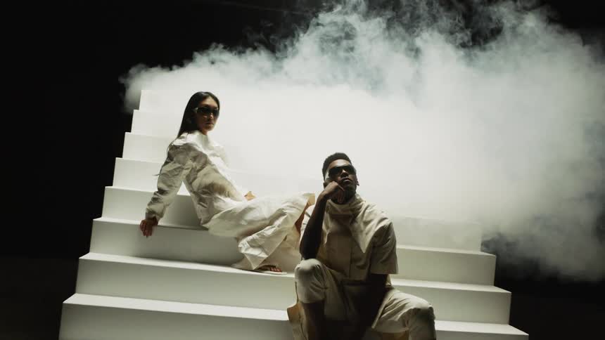 A man and woman sit on a freestanding set of stairs while posing for a fashion photo shoot