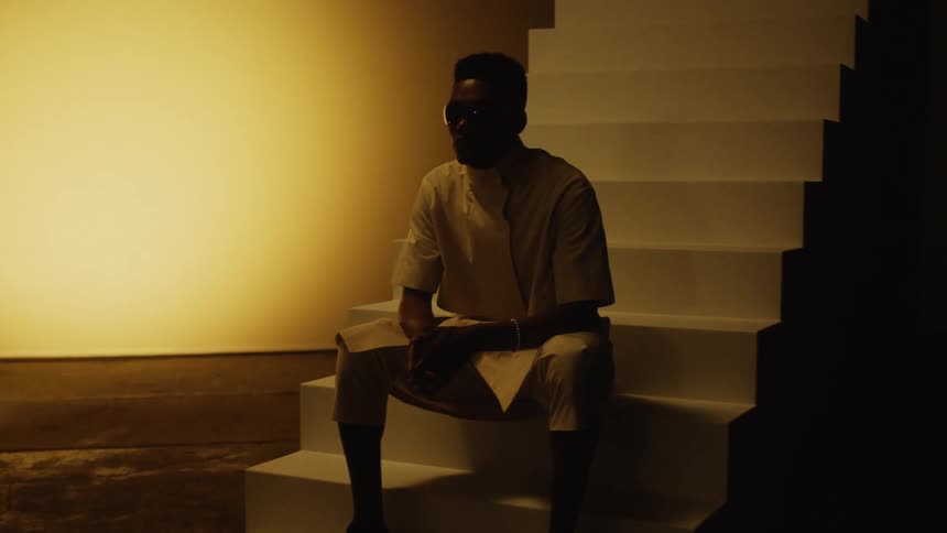 Woman sits in silhouette on one set of stairs while man sits on second set of stairs on shadowed stage with hands pressed together as light forms circle on wall