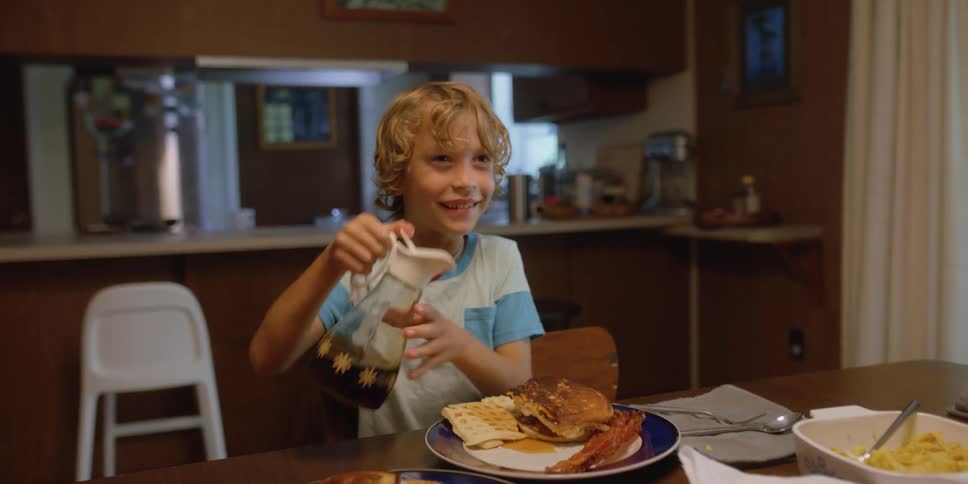 A boy sitting on a table pouring syrup onto a plate with pancakes and waffles
