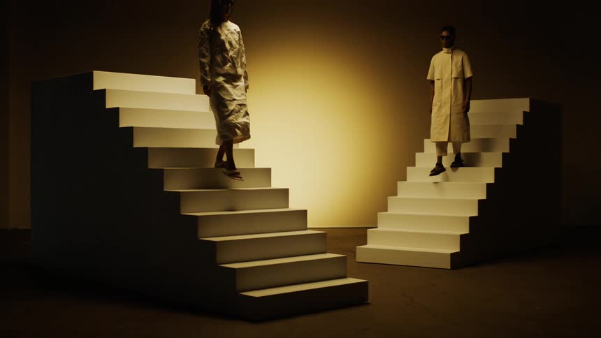 Two people modeling fashionable attire slowly descend two freestanding stairways