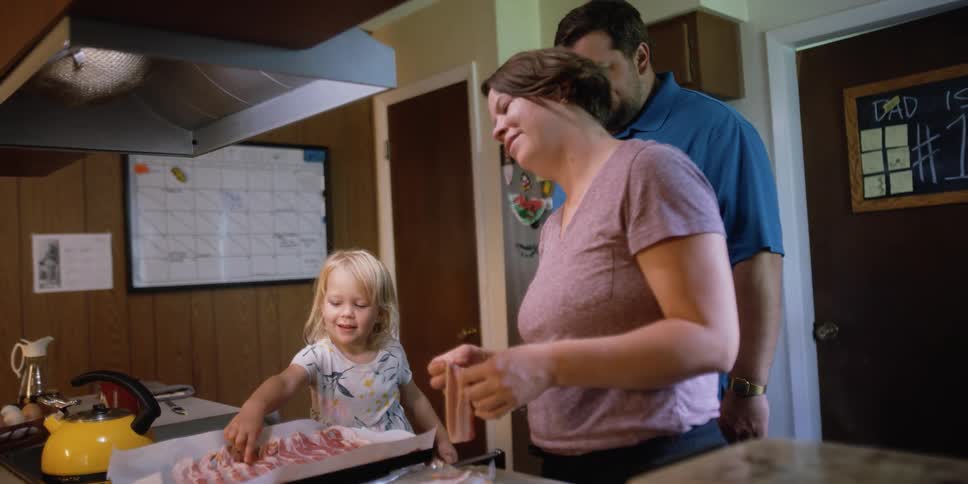 Mother and father watch as their daughter counts the bacon slices that are on wax paper in a baking pan