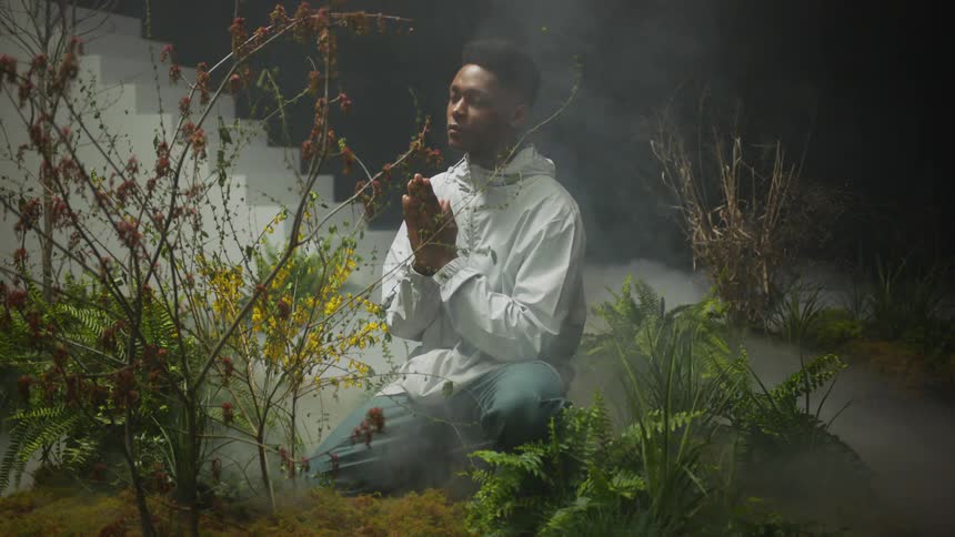 A young man prays while kneeling before vegetation in a dark and misty expanse