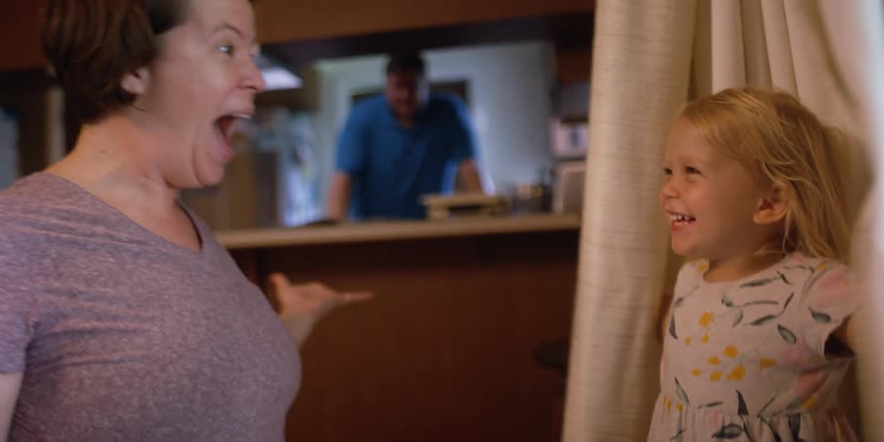 A little girl in a room hiding in curtains and revealing herself to a woman with a man in the background