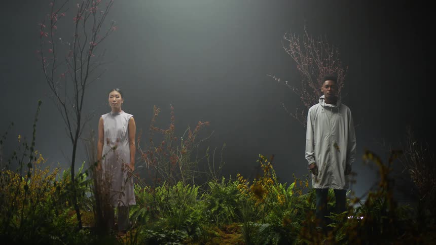 A man and woman stand near one another in a patch of vegetation in an otherwise empty area shrouded in darkness and fog