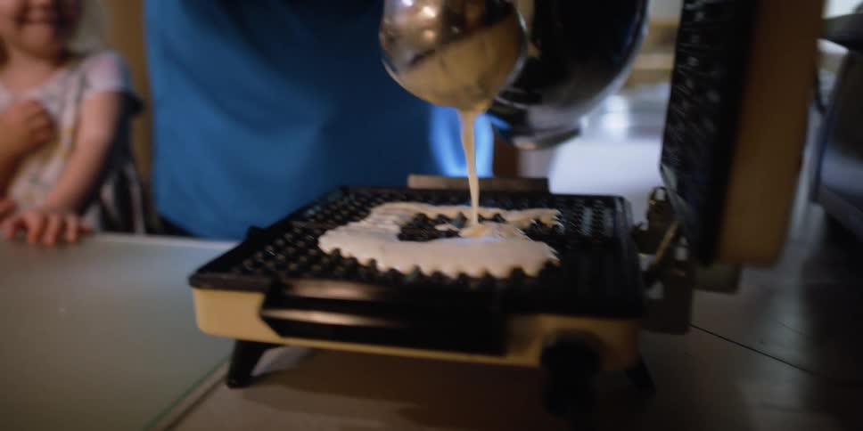 A man in a kitchen pouring pancake batter on a waffle iron with a girl and a woman in the background
