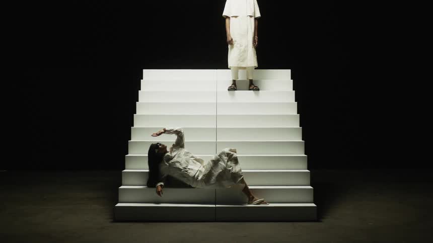 Woman reclines on white stairs while another person stands near top of movable stairs both wearing white in dimly lit area