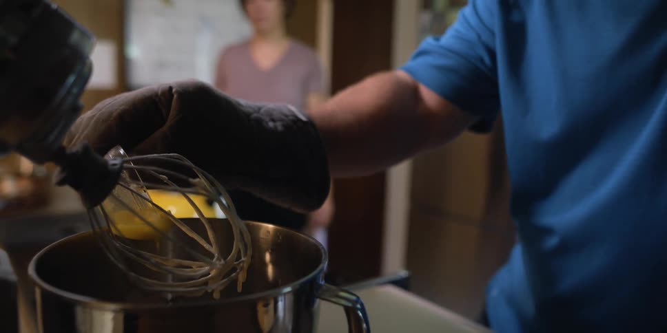 A man pours an ingredient in to a bowl as he cooks while a woman stands in the background talking to him