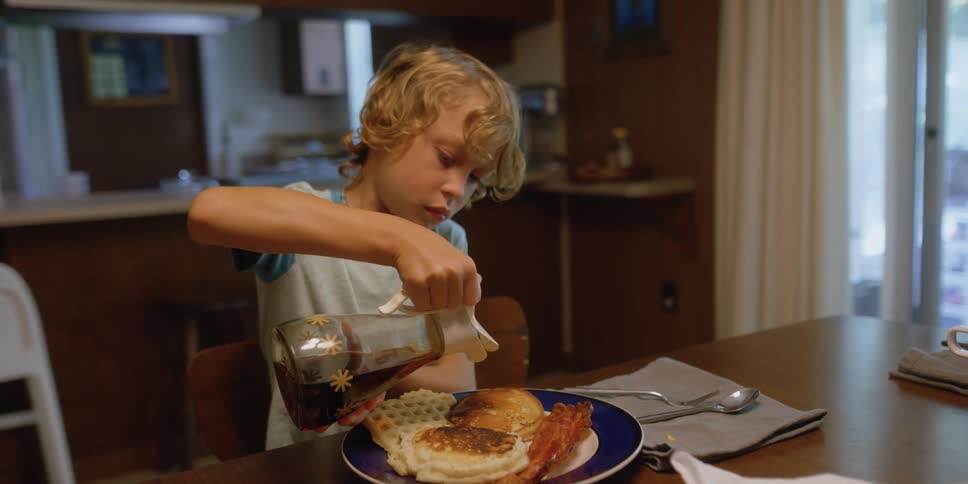 A young boy pours syrup on a plate of breakfast food at home