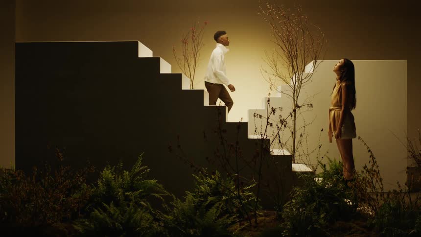 A man and a woman on a stage with staircases and plants with a wall in the background