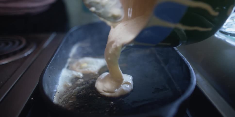 A woman pours batter onto a skillet as a child behind her watches