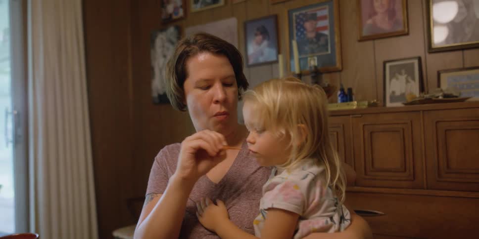 A woman takes a bite of food and offers it to the girl sitting on her lap who takes a bite