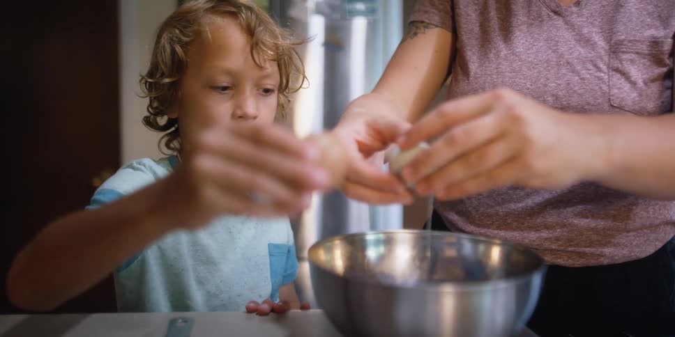 A male child and a woman crack eggs into a silver mixing bowl on a counter top