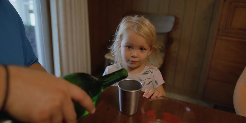 A child walks over to her parents with a cup and her father pours her a water from a bottle at home in a dining room