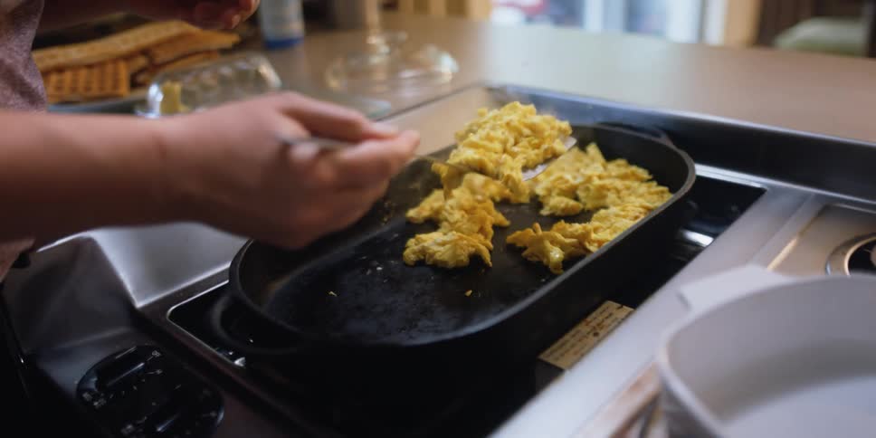 A woman in a kitchen cooking eggs on a skillet and using a spatula to place the eggs in a container