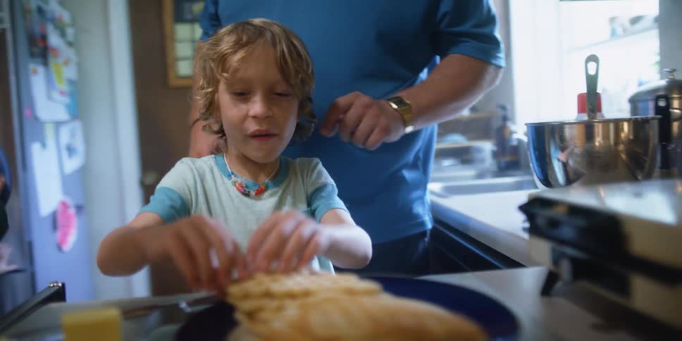 A woman stirs batter and pours it in a pan as her son comes and grabs a waffle and takes a bite and his dad taps his shoulder
