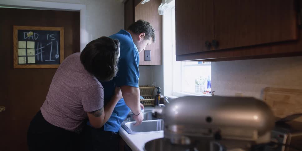 A woman is hugging a man while hes washing dishes
