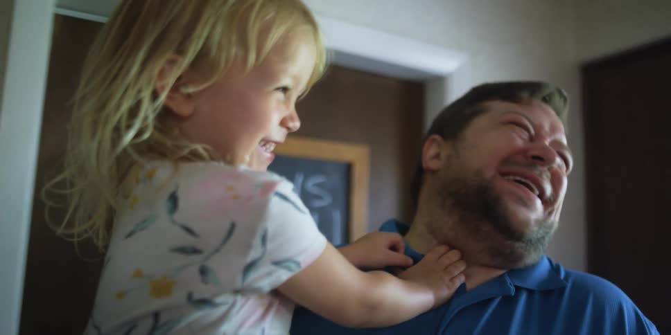 A man carrying a young girl in his arms kisses her on the cheek and laughs when she turns to look at him