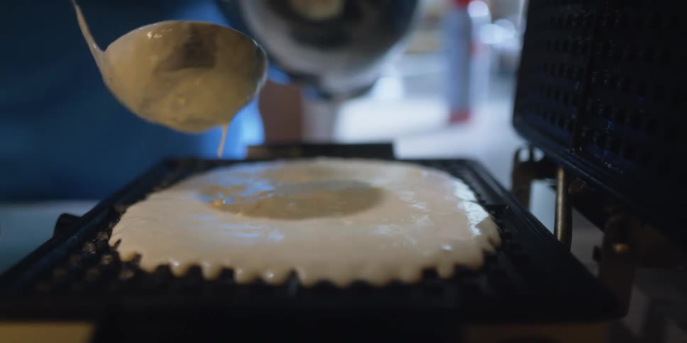 A person in a kitchen spreading pancake batter on a waffle iron with a spoon while holding a mixing bowl