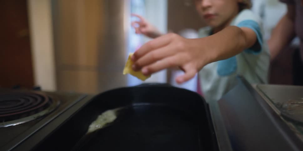 A boy in the kitchen putting butter on a skillet on a stove