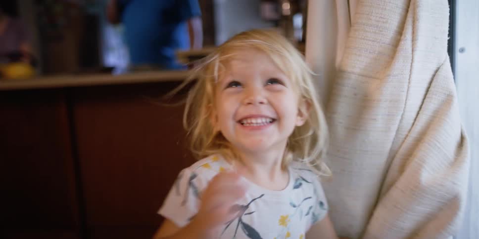 A little girl in a room wrapping herself in a curtain with a stool and a counter in the background