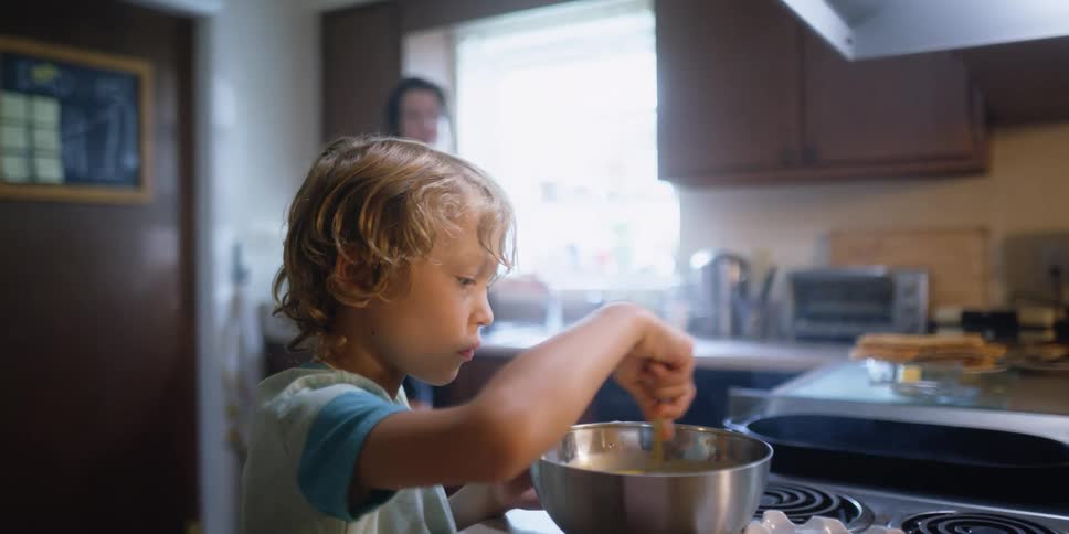 A woman pours a small amount of liquid into a bowl that a young boy is stirring in their home kitchen