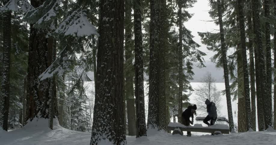 Two people walk through snowy woods and clear off a bench