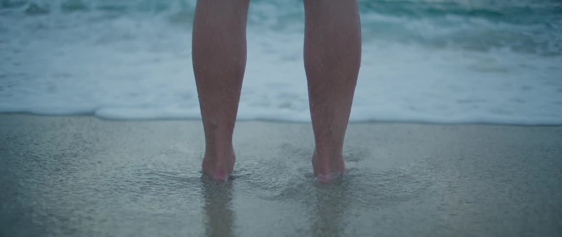 A person stands in front of a body of water as the water laps their feet and ankles