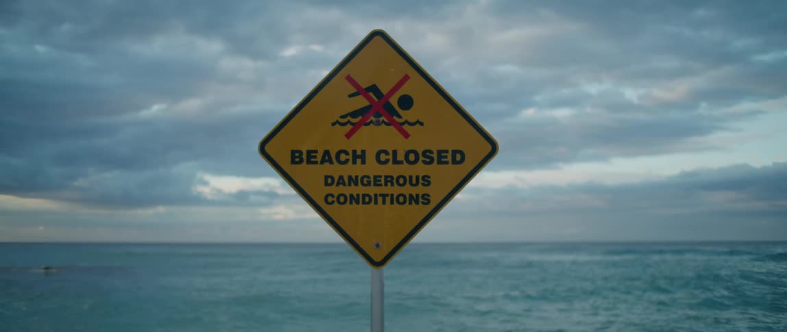 Sign stating the beach is closed due to dangerous conditions with the ocean in the distance under a cloudy sky
