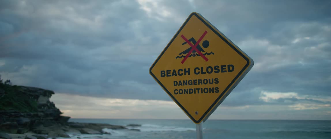 Sign stating the beach is closed due to dangerous conditions with the ocean in the distance under a cloudy sky