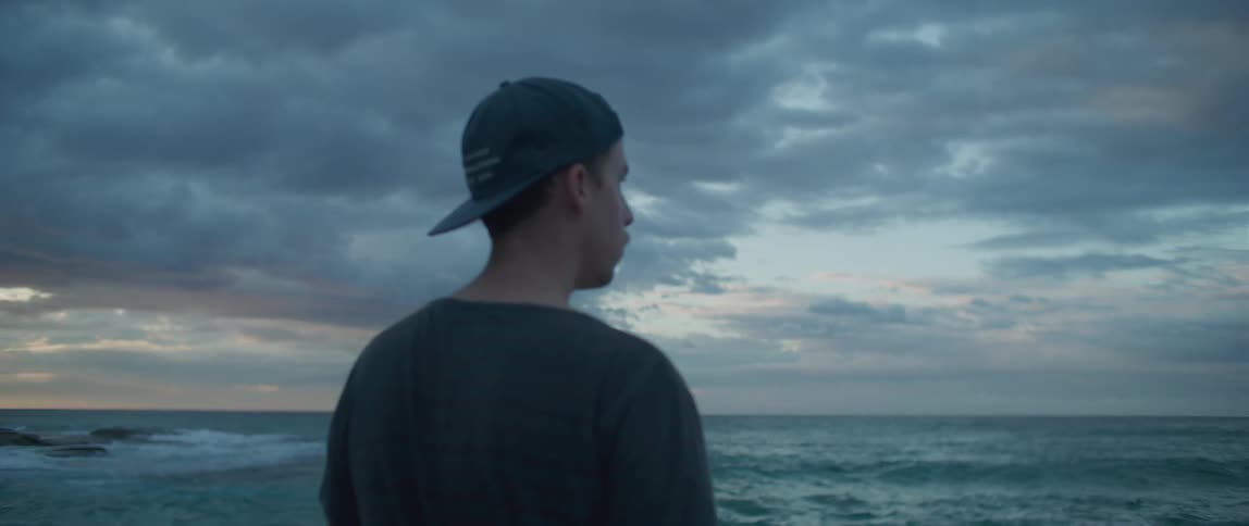 A man with a hat standing near the coast with the ocean sky and clouds in the background