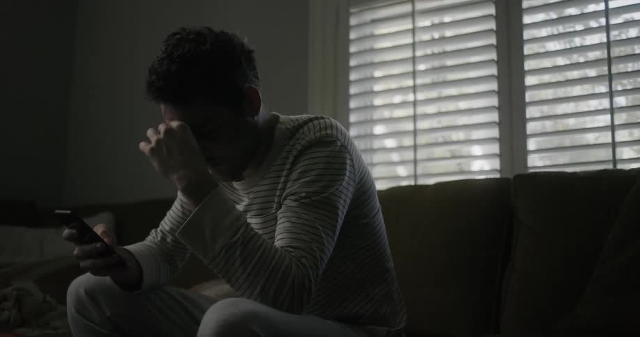 A man in a room sitting on a couch looking at a smart phone with a wall and window in the background